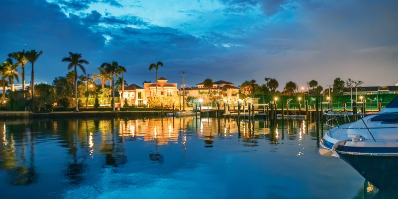 Boca Raton waterfront homes with palm trees and docks are illuminated at dusk, reflecting in calm water under a partly cloudy sky.