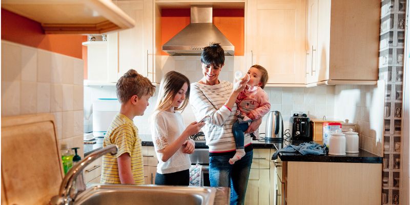 A woman stands in a kitchen holding a young child while two older children stand nearby, one looking at a phone.