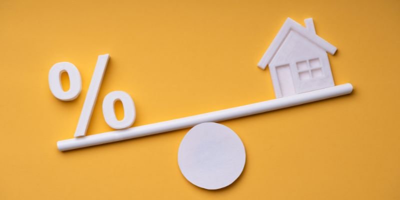 A white percentage sign and a small house icon are balanced on opposite ends of a seesaw against a yellow background, visually representing mortgage rates and home ownership.