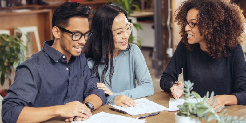 Three people sit at a table with documents, engaged in conversation. All are smiling and appear to be in a casual office setting.