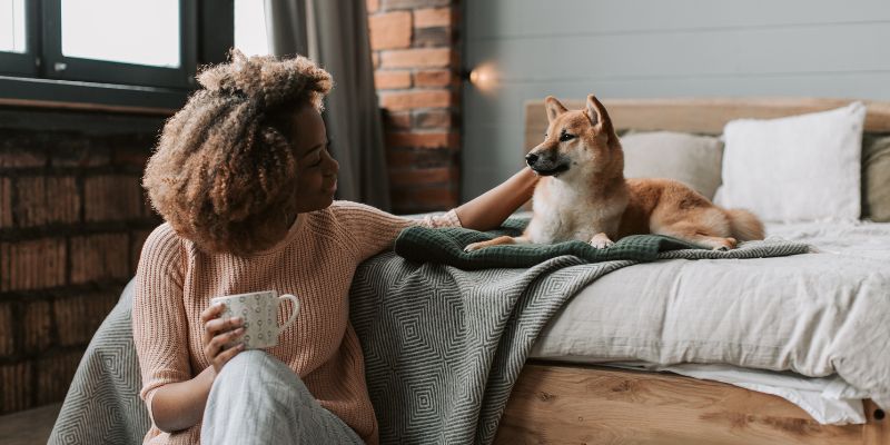 A person with curly hair sits on the edge of a bed holding a mug, while a Shiba Inu dog lies on the bed.