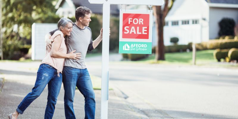 Couple standing outside their new home by the "For Sale" sign