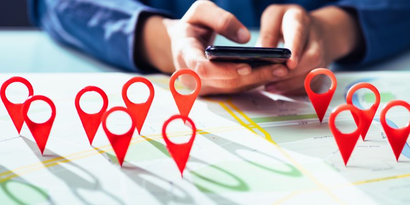 A person uses a smartphone behind a map with multiple red location pins placed on it, suggesting the concept of searching for a home.