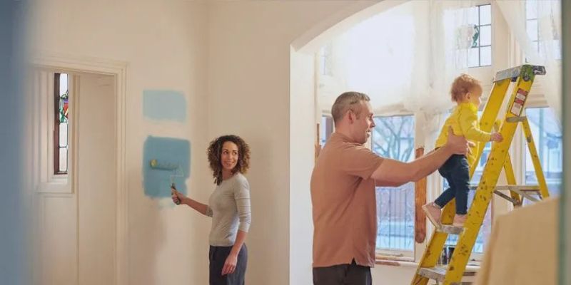 A woman paints a patch of blue on a white wall while a man helps a young child climb a yellow ladder in a bright room with large windows.